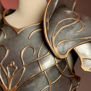 May include: Close-up of a gray and gold fantasy armor piece. The armor features intricate gold filigree designs and is displayed on a mannequin. The shoulder and chest plates are visible, showcasing the detailed craftsmanship.