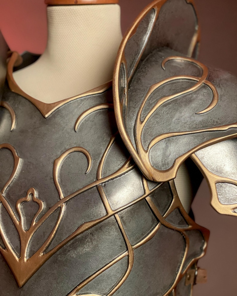 May include: Close-up of a gray and gold fantasy armor piece. The armor features intricate gold filigree designs and is displayed on a mannequin. The shoulder and chest plates are visible, showcasing the detailed craftsmanship.