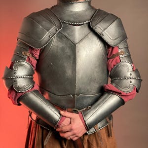 May include: A silver-colored medieval armor set, featuring a breastplate, shoulder guards, and arm protection. The armor is detailed with rivets and rope-like trim. Red fabric is visible beneath the arm protection. The armor is worn with brown pants and a belt.