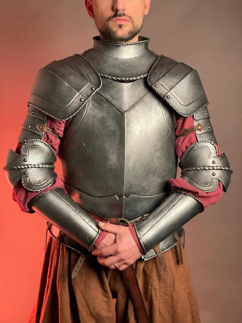 May include: A silver-colored medieval armor set, featuring a breastplate, shoulder guards, and arm protection. The armor is detailed with rivets and rope-like trim. Red fabric is visible beneath the arm protection. The armor is worn with brown pants and a belt.