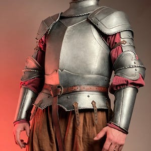May include: A silver-colored medieval armor set, including a chest plate, shoulder guards, and arm protection. The armor is worn over a red and brown outfit, with a brown leather belt and accents. The design is detailed and appears to be handcrafted.