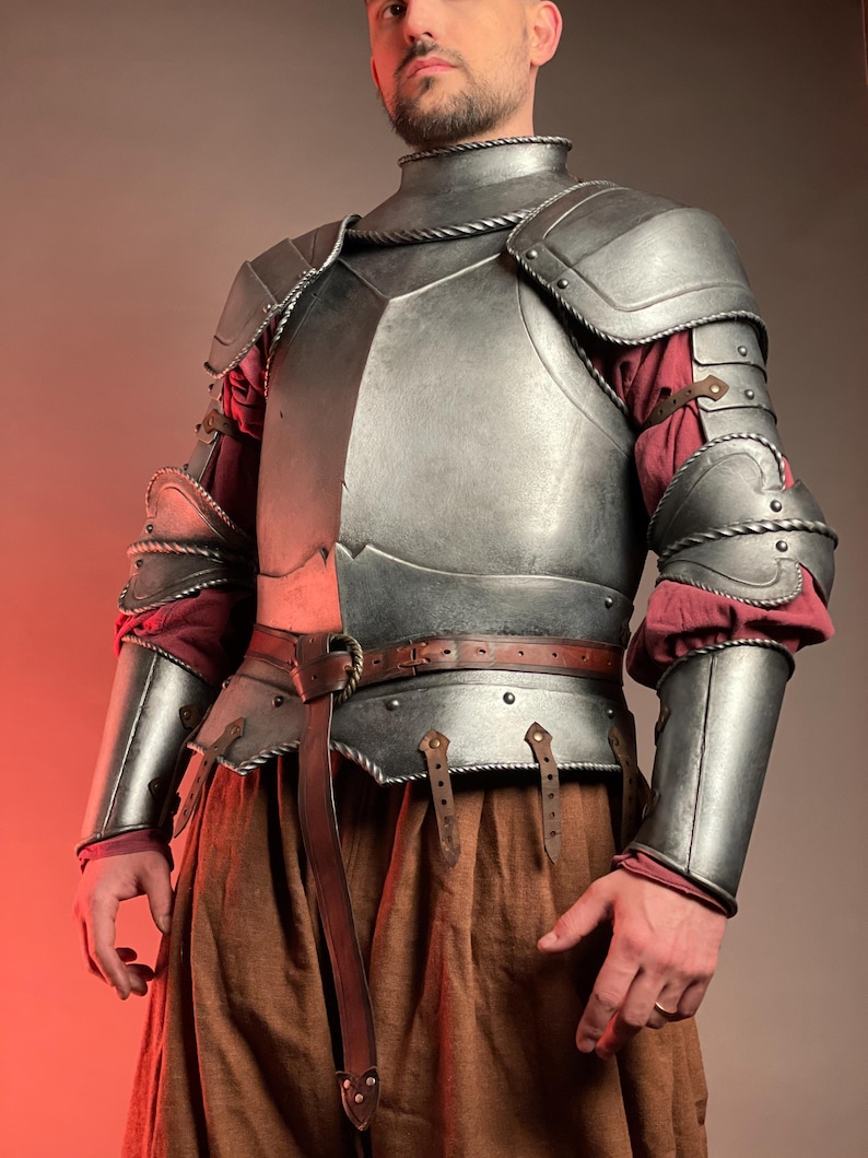 May include: A silver-colored medieval armor set, including a chest plate, shoulder guards, and arm protection. The armor is worn over a red and brown outfit, with a brown leather belt and accents. The design is detailed and appears to be handcrafted.