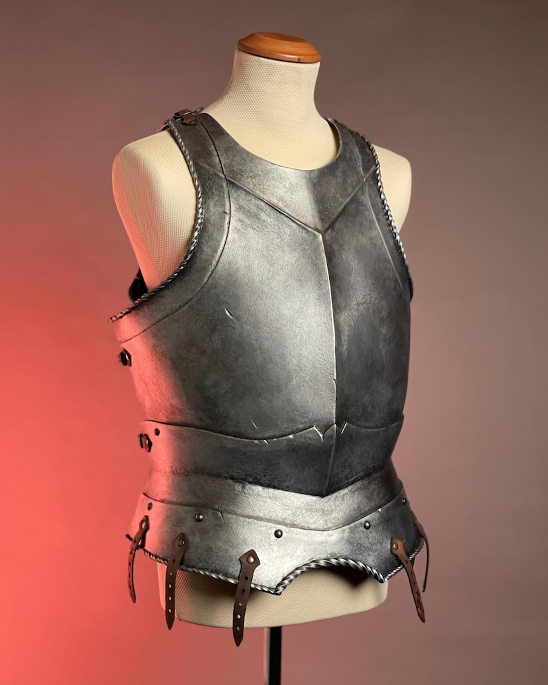 May include: A silver-colored breastplate, displayed on a mannequin. The armor features a detailed design with visible rivets, leather straps, and a decorative trim. The chest piece is designed to protect the torso and is likely part of a medieval or fantasy costume.