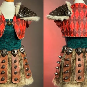 May include: A costume comprising a red and brown diamond-patterned vest with fur trim and shoulder armour. The outfit includes a green top, a fur-trimmed skirt with leather accents, and metal embellishments. The costume is displayed on a mannequin.
