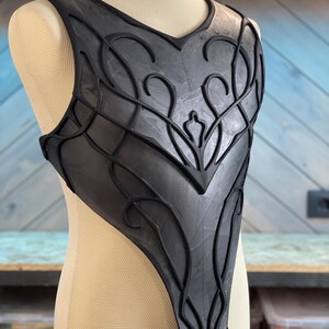 May include: Black chest armor with intricate, raised floral and geometric designs. The armor is displayed on a mannequin and appears to be made of a flexible material. The design features a V-neck and a fitted shape, suggesting a fantasy or cosplay theme.