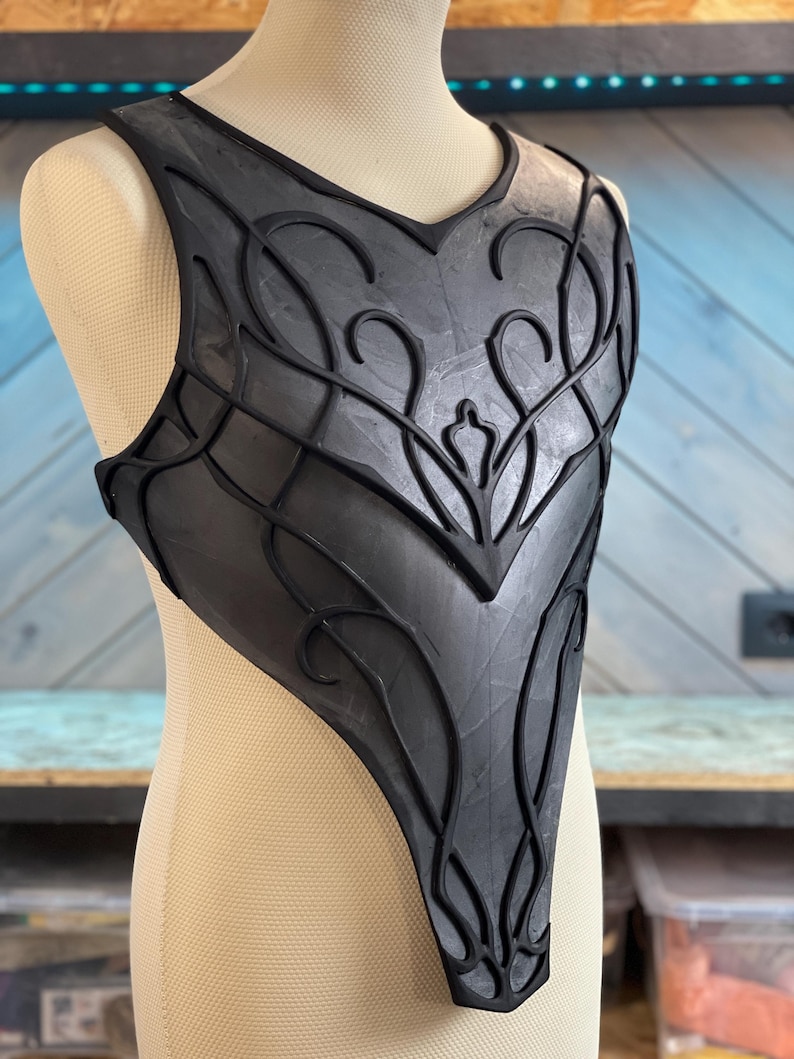 May include: Black chest armor with intricate, raised floral and geometric designs. The armor is displayed on a mannequin and appears to be made of a flexible material. The design features a V-neck and a fitted shape, suggesting a fantasy or cosplay theme.