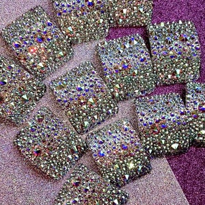 May include: A close-up of a variety of silver and iridescent rhinestones arranged in a pattern on a purple background. The rhinestones are all different shapes and sizes, and they sparkle in the light.
