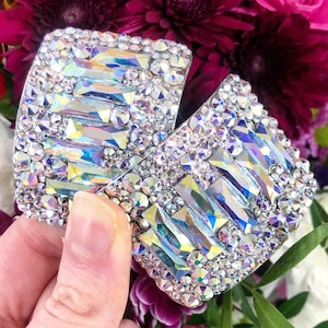 May include: A pair of silver-toned earrings with a geometric design made of iridescent rhinestones. The earrings are shaped like a rectangle with rounded corners.