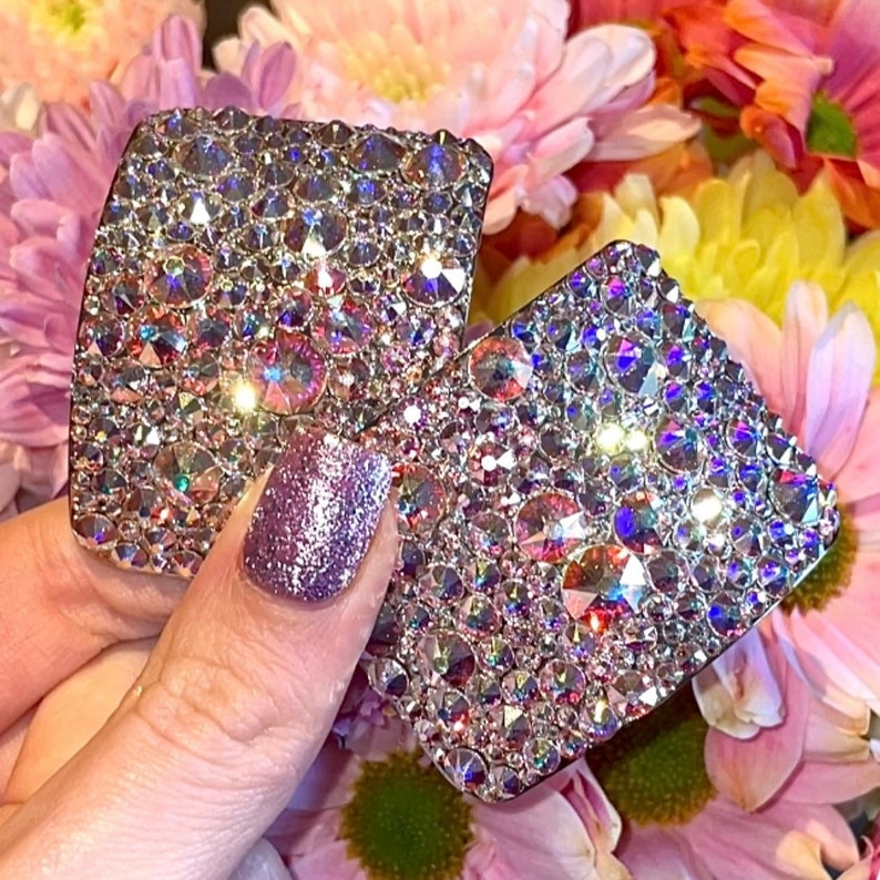 May include: Two square-shaped hair clips with a sparkly, iridescent finish. The clips are made of rhinestones and have a silver backing.