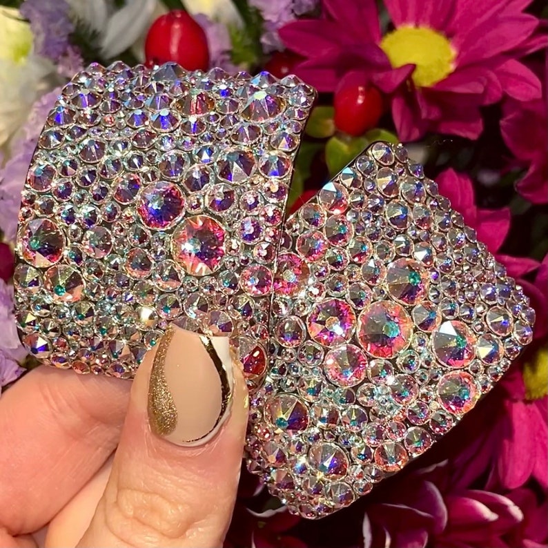 May include: A pair of square-shaped earrings with a silver base covered in iridescent rhinestones. The rhinestones are in shades of pink, purple, and blue.