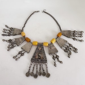 Könnte beinhalten: Eine silberfarbene Halskette mit geometrischem Design, mit bernsteinfarbenen Perlen und dekorativen Metallarbeiten. Die Halskette hat eine Kette und mehrere anhängerartige Elemente mit hängenden Akzenten, die ein detailliertes und verziertes Aussehen erzeugen.