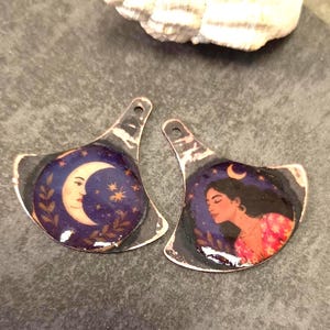 Peut inclure: Deux pendentifs en métal en forme d'éventail avec une finition en cuivre oxydé foncé. L'un présente un croissant de lune avec un visage sur un ciel étoilé. L'autre montre une personne aux cheveux foncés portant un haut floral sous une lune.