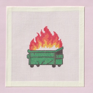 May include: A square needlepoint canvas featuring a cross-stitch design of a green dumpster on fire. The dumpster is rendered in shades of green with black outlines, while the flames above are depicted in vibrant layers of yellow, orange, and red.