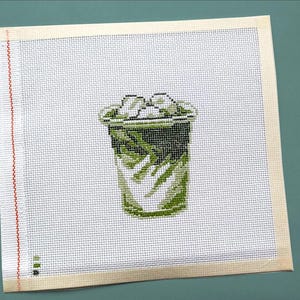 May include: Needlepoint canvas featuring a green and black iced beverage with ice cubes. The design is stitched on a white canvas with a beige border. The canvas has a red stitch line on the left side.