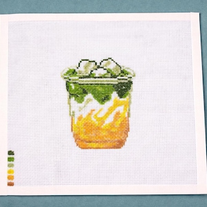 May include: Cross-stitch design of a refreshing beverage in a clear glass. The drink features layers of green, white, and yellow, with ice cubes on top. The design is on a white canvas with a color palette on the left.
