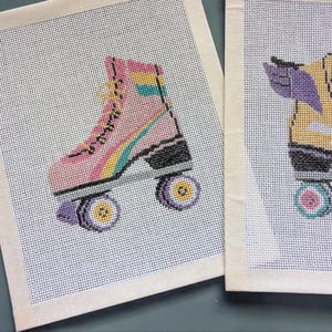 May include: Two needlepoint canvases featuring roller skates. The left canvas shows a pink skate with a rainbow stripe, yellow laces, and purple wheels. The right canvas displays a yellow skate with purple wings, black laces, and teal wheels.