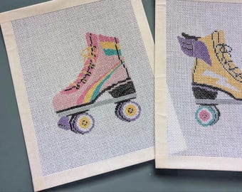 May include: Two needlepoint canvases featuring roller skates. The left canvas shows a pink skate with a rainbow stripe, yellow laces, and purple wheels. The right canvas displays a yellow skate with purple wings, black laces, and teal wheels.