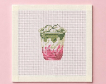 May include: A square needlepoint canvas featuring a cross-stitch design of a layered iced beverage in a clear plastic cup. The drink shows distinct layers of dark green matcha, creamy white milk, and a vibrant pink syrup base with ice cubes.