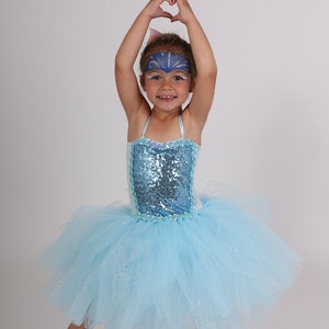 May include: A young child in a light blue tutu dress with a sequined bodice and spaghetti straps. The child has face paint and is making a heart shape with their hands. The tutu has a layered, fluffy design.