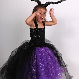 Könnte beinhalten: Ein Halloween-Kostüm für Kinder mit einem schwarzen Pailletten-Oberteil und einem mehrlagigen Tüllrock in Schwarz und Lila. Das Kostüm enthält ein schwarzes Horn-Stirnband. Das Kind trägt das Kostüm und hat die Hände oben.