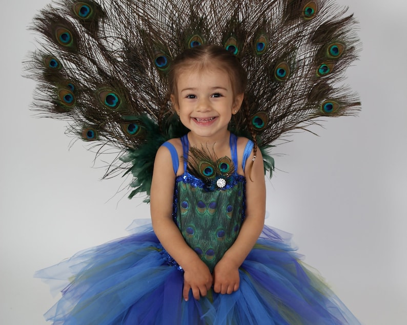 Une jeune fille porte un costume de paon. La robe présente un corsage bleu avec un motif de plumes de paon et une jupe en tulle bleue, verte et violette superposée. Un grand éventail de plumes de paon orne le dos.