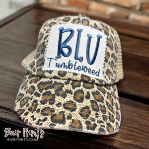 May include: A leopard print trucker hat with a blue and white patch that says "BLU" and "Tumbleweed".