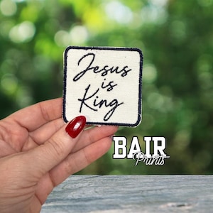 May include: A square patch featuring the text "Jesus is King" in black script on a cream-coloured background, with a navy blue border. The patch is held in a hand with red nail polish. The brand name "BAIR Prints" is displayed below.