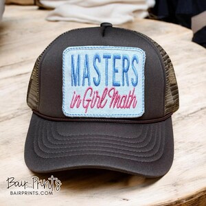 May include: A brown trucker hat with a mesh back and a curved brim. The front patch reads "MASTERS in Girl Math" in blue and pink embroidery. The hat has a dark brown crown and a brown brim.