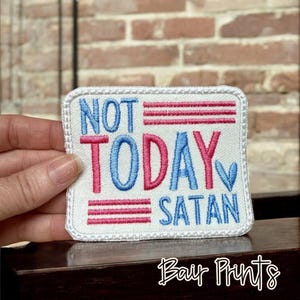 May include: Embroidered patch with the words "NOT TODAY SATAN" in blue and pink lettering. The patch has a white border and red and white stripes. The patch is held by a hand.