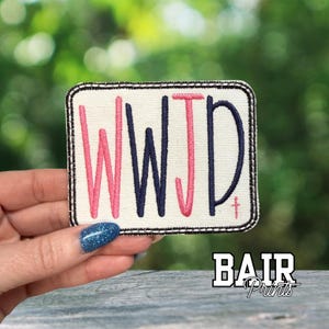 May include: A rectangular patch with the letters "WWJD" in pink and navy blue, stitched on a white background with a black border. The patch also features a small cross and the text "BAIR Prints".
