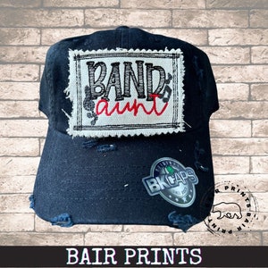 May include: Black distressed baseball cap with a white patch that says "Band aunt" in black and red lettering. The patch has a black border and a distressed look. The cap has a black adjustable strap and a silver metal tag that says "Original BK Caps Since 2003".
