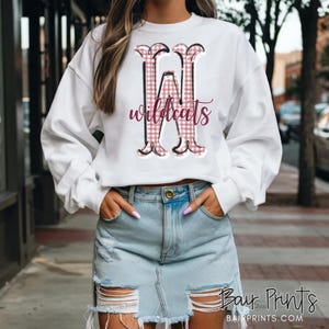 May include: White sweatshirt with a large, stylized letter "W" in a pink and black plaid pattern, with the word "wildcats" in a cursive font. The sweatshirt has long sleeves.