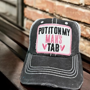 May include: A black distressed baseball cap with a white and pink patch that says "Put it on my man's tab".