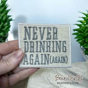 May include: A beige fabric patch with white stitching around the edges. The patch has the text "NEVER DRINKING AGAIN (AGAIN)" embroidered in grey thread.