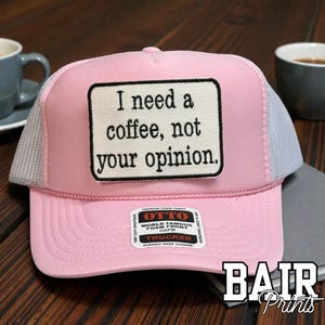 May include: Pink trucker hat with a white mesh back and a rectangular patch that reads "I need a coffee, not your opinion." The hat has a curved brim and a snap closure. The brand name "BAIR Prints" is visible.