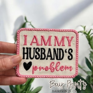 May include: Embroidered patch with the text "I AM MY HUSBAND'S problem" in pink and black, with a black heart. The patch has a pink border and is on a cream-colored background.
