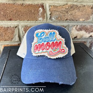 May include: A blue and white baseball cap with a distressed look. The cap has a white mesh back and a blue front with a red, yellow, and blue embroidered patch that says "Ball Mom".