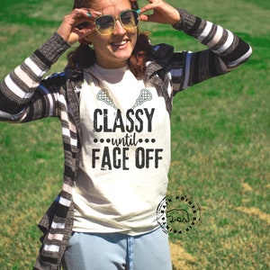 May include: Cream-colored t-shirt with the text "Classy until Face Off" in black, along with lacrosse stick graphics. The shirt is worn with a striped cardigan and sunglasses. The image is taken outdoors on a sunny day.
