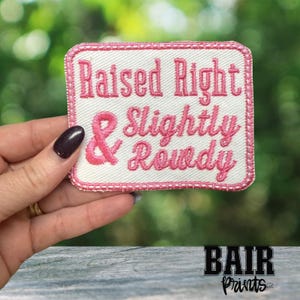 May include: A rectangular patch with a white background and pink embroidered text that reads "Raised Right & Slightly Rowdy." The patch has a pink border and a pink ribbon symbol. The patch is held in a hand.