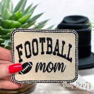 May include: A beige embroidered patch with black stitching. The patch features the text "FOOTBALL mom" with a black and white football graphic.