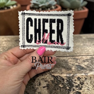 May include: A rectangular fabric patch with a raw, frayed edge featuring a black embroidered border and the word CHEER in bold black letters. The word Mom is embroidered in pink cursive script across the bottom of the black text.