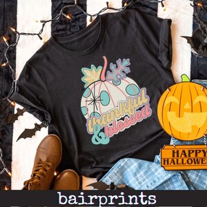 以下が含まれることがあります： カラフルなカボチャのデザインと「thankful & blessed」の文字が入った黒いTシャツ。カボチャは白いベースに青い水玉模様とパステルカラーのアクセント。装飾用のカボチャと「Happy Halloween」のサインも表示されています。