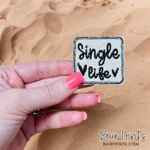May include: A white fabric patch with black embroidery that reads "Single Life" with a heart on each side of the word "life".