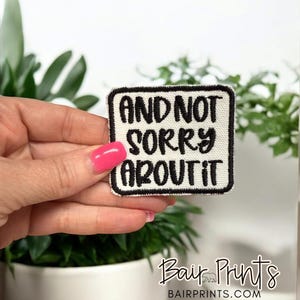 May include: A square patch with the text "AND NOT SORRY ABOUT IT" in black, outlined in black, on a white background. The patch is held in a hand with pink nail polish, with green plants in the background. The patch is from Bair Prints.