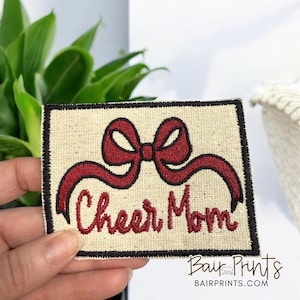 May include: A white fabric patch with a black border, featuring a red embroidered bow and the text "Cheer Mom".