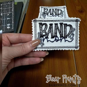 May include: Embroidered white fabric patch with the words "BAND mom" in black script. The patch has a black border and decorative musical notes. The patch is held in a hand.