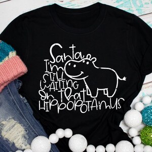May include: Black t-shirt with white text and a hippo graphic. The text reads "Santa I'm still waiting on that hippopotamus." A pink, blue, and white knit hat and denim jeans are next to the shirt.