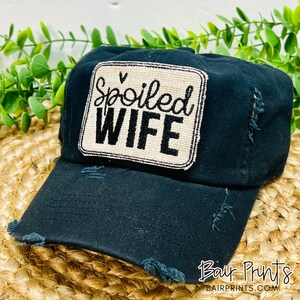 May include: Black distressed baseball cap with a beige patch that says "Spoiled Wife" in black cursive lettering.