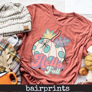 May include: Rust-colored t-shirt featuring a whimsical pumpkin design with the words "Hello Fall" in pastel colors. The pumpkin has blue polka dots and is surrounded by leaves. A knit beanie and scarf are next to the shirt.