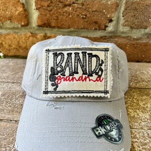 May include: Gray distressed baseball cap with a white patch that says "Band Grandma" in black lettering with a red musical note. The cap has a black and silver patch that says "Original BK Caps Since 2007".